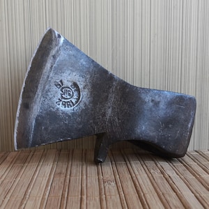 May include: A weathered metal axe head with a wide, curved blade and a dark, oxidized finish. A circular stamp is visible on the side, featuring the text "BK" and "1892" arranged around a central symbol. The tool has a distinct, heavy appearance.