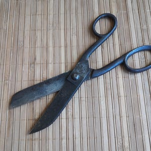 Puede incluir: Grandes tijeras metálicas antiguas con mangos y hojas negras. Las tijeras están abiertas, revelando las afiladas hojas. Las tijeras descansan sobre una estera de bambú marrón claro.
