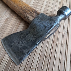 Könnte beinhalten: Eine alte Axt mit einem Holzgriff und einem dunklen, verwitterten Metallkopf. Der Axtkopf hat einen Hammer auf der gegenüberliegenden Seite der Klinge. Der Holzgriff weist Gebrauchsspuren auf. Die Axt liegt auf einer hellbraunen, strukturierten Oberfläche.
