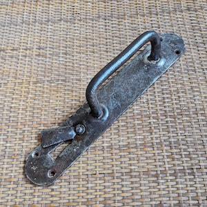 Puede incluir: Un tirador de puerta de metal antiguo y desgastado con un asa curvada y una placa trasera rectangular. El tirador y la placa trasera tienen un acabado oscuro y envejecido, mostrando signos de desgaste. El tirador está montado sobre una superficie tejida.