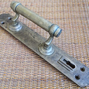 May include: A tarnished brass door handle set featuring a long rectangular backplate with rounded ends, a cylindrical horizontal grip, and a keyhole escutcheon. The metal surface shows signs of age and wear with visible screw holes for mounting.
