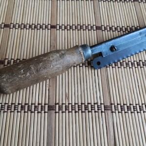 May include: A vintage saw with a wooden handle and a metal blade. The blade has a serrated edge and a small metal loop at the end. The saw is lying on a woven bamboo mat.