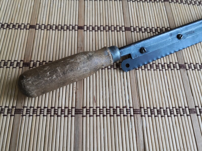 May include: A vintage saw with a wooden handle and a metal blade. The blade has a serrated edge and a small metal loop at the end. The saw is lying on a woven bamboo mat.