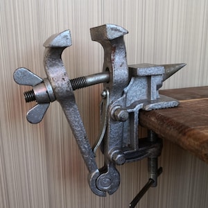 May include: A silver-colored metal bench vise with a textured finish, featuring a threaded screw mechanism with a wing nut, a stationary jaw, a movable jaw, and an integrated anvil horn, secured to a wooden surface with a bottom clamp screw.