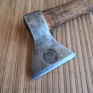 May include: A metal axe head with a wooden handle featuring a circular stamp on the blade that reads ZIG and 1965. The steel head shows signs of wear and oxidation, while the wooden handle has a natural brown grain and a smooth, polished finish.