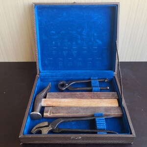 May include: A brown storage case with a vibrant blue velvet interior containing a set of tools. The set includes a hammer with a wooden handle, two pairs of dark metal pincers, and two rectangular wooden blocks arranged neatly inside the case.