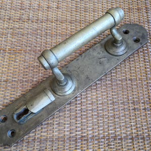 Könnte beinhalten: Ein Türgriff-Set aus Messing mit einer langen, rechteckigen Grundplatte mit abgerundeten Enden, einem zylindrischen horizontalen Griff und einer Schlüssellochöffnung. Die Metalloberfläche zeigt eine verwitterte Patina mit Gebrauchsspuren.