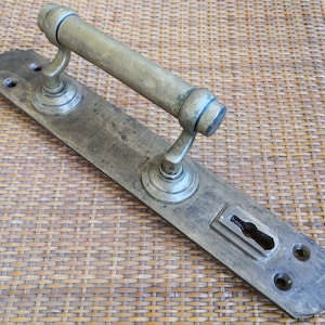 Pode incluir: Maçaneta de porta antiga de latão com uma placa posterior retangular longa e uma fechadura. A maçaneta tem uma forma redonda e cilíndrica e um design decorativo.