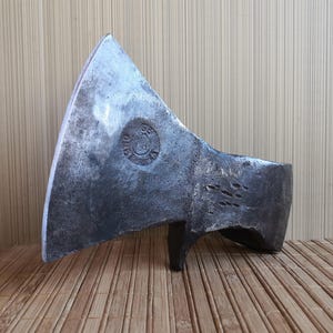 以下が含まれることがあります： 風化したダークグレーのパティーナ仕上げが施された、手鍛造のスチール製斧頭。刃は幅広く湾曲した刃先を持ち、金属表面には円形のメーカー刻印が打たれている。斧の背部分は厚みのある長方形で、ハンマーの打痕がはっきりと確認できる。