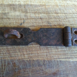 May include: A rusty metal latch with a hinged design. The latch has a loop on one end and two holes on the other end.