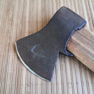 Könnte beinhalten: Ein Handbeil mit einem dunklen, verwitterten Metallkopf, der ein schwach eingeprägtes Logo und eine geschwungene, geschärfte Klinge aufweist. Der Kopf ist fest mit einem hellen Naturholzstiel mit sichtbarer Maserung verbunden.