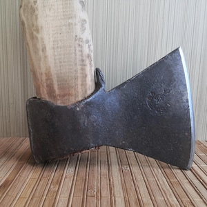 May include: A hand axe with a weathered, dark gray metal head featuring a stamped circular mark and a light-colored, textured wooden handle. The metal blade shows signs of age and use, with a sharp, polished edge contrasting against the dark iron.