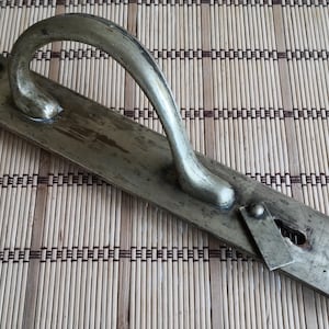 Op de afbeelding: Een vintage metalen deurklink met een gebogen handvat en een rechthoekige achterplaat. De klink en achterplaat hebben een aangetaste, verouderde uitstraling. De klink is ongeveer 15 cm lang.