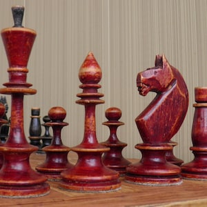 May include: A set of vintage wooden chess pieces in shades of red and black. The pieces include a king, bishop, knight, and pawns, all displayed on a wooden surface. The chess pieces are intricately carved and show signs of age.