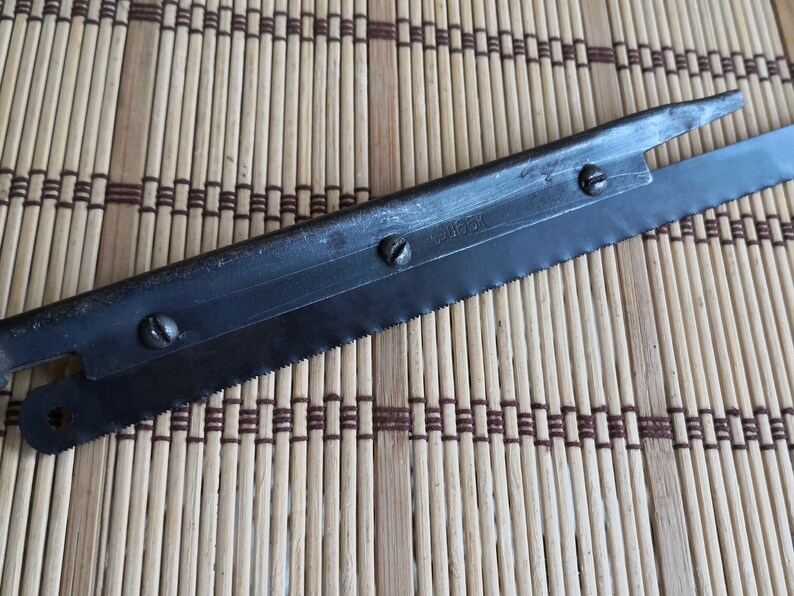 May include: A black metal saw blade with a serrated edge and a small handle. The blade has four screws and the text "2165K" printed on it.