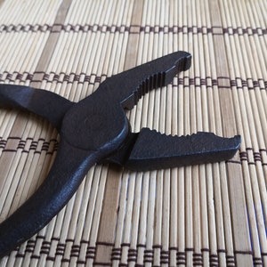 May include: Black metal pliers with a serrated jaw and a smooth handle. The pliers are lying on a woven bamboo mat.