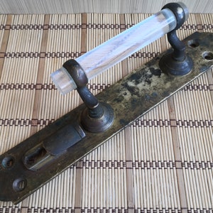 May include: A brass door handle with a clear glass pull. The handle has a keyhole and is mounted on a rectangular plate.