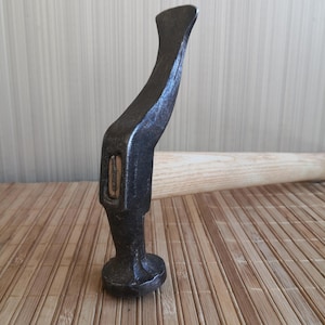 May include: A shoemaker's hammer featuring a dark, weathered metal head with a curved, elongated claw and a round, flat striking face. The head is securely attached to a light-colored, smooth wooden handle that extends horizontally from the metal.