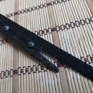 May include: A black metal saw blade with a serrated edge and a small metal handle with four screws. The saw blade is lying on a brown and white bamboo mat.