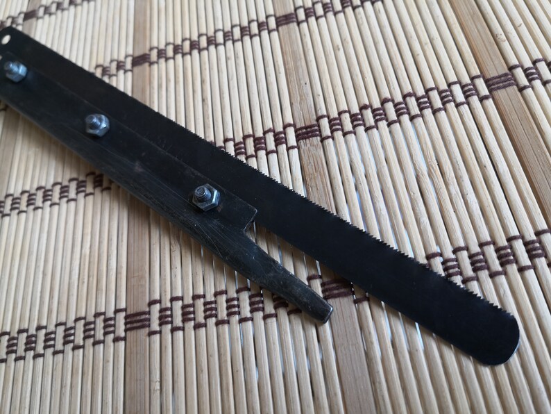 May include: A black metal saw blade with a serrated edge and a small metal handle with four screws. The saw blade is lying on a brown and white bamboo mat.