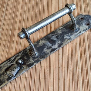 May include: A vintage metal door handle with a silver-colored bar and a black metal plate. The handle has a worn, distressed look.