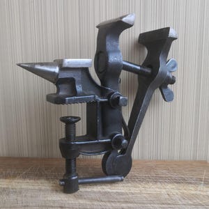 May include: A black cast iron combination tool featuring a small anvil with a pointed horn and a flat top, integrated with a bench vise. The tool includes a threaded screw clamp mechanism for mounting and a wing nut for adjusting the vise jaws.