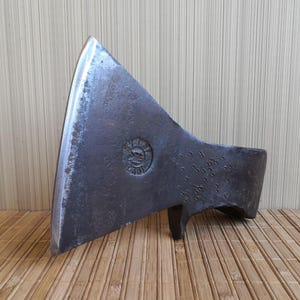 May include: A vintage axe head with a worn, metallic finish. The axe head features a sharp, curved blade and a circular emblem. The axe is displayed on a bamboo surface, suggesting a rustic or antique theme.