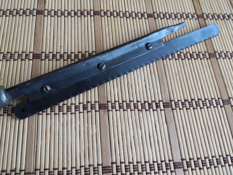 May include: A close-up of a metal saw blade with a handle. The blade has a serrated edge and a small hole near the handle. The blade is marked with the number "2467".