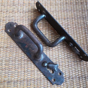 Może przedstawiać: Dwie antyczne metalowe klamki do drzwi o rustykalnym wykończeniu. Jedna klamka ma prosty design pętli, podczas gdy druga ma bardziej ozdobny design z ozdobnym pokrętłem.