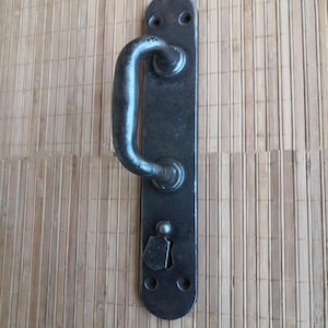 Może przedstawiać: Czarny metalowy klamka do drzwi z prostokątną płytką o zaokrąglonych krawędziach i uchwytem w kształcie litery D. Klamka ma dziurkę od klucza i dwa małe śruby na płytce.