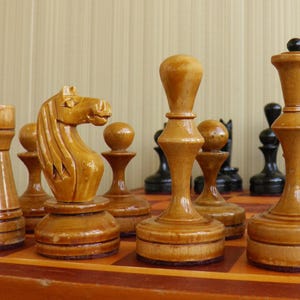 May include: A set of polished light brown wooden chess pieces including a knight, a rook, a bishop, and a queen arranged on a wooden chessboard. In the background, several dark black chess pieces are visible, creating a contrast in color.