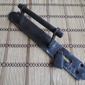 May include: A rustic, dark metal door pull with a horizontal handle and a long, rectangular backplate. The handle is cylindrical, and the backplate has decorative cutouts and mounting holes. The hardware has a weathered appearance.