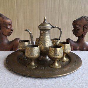 May include: A brass serving set featuring a teapot, six cups, and a tray. The set is adorned with elaborate floral designs. The set is presented on a white tablecloth alongside two wooden sculptures of human heads.