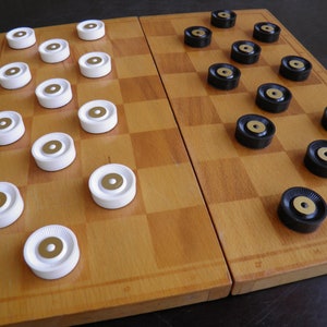 Puede incluir: Un juego de damas de madera con fichas negras y blancas. Las fichas están dispuestas en un patrón en el tablero. El tablero está hecho de madera y tiene un acabado marrón claro.