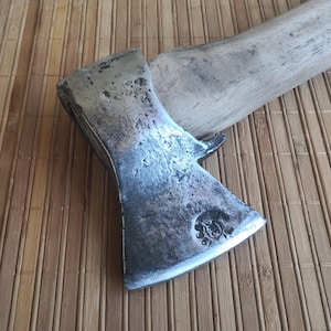 May include: A hand axe featuring a weathered, dark metal head with a textured, pitted surface and a stamped circular maker's mark near the sharpened edge. The axe is attached to a light-colored, unfinished wooden handle with a visible grain.