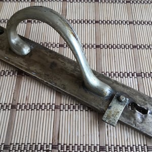 Może przedstawiać: Antyczny mosiężny uchwyt do drzwi z zakrzywionym uchwytem i prostokątną płytką. Uchwyt ma otwór na klucz i otwory montażowe, wykazujące ślady zużycia. Okucia są w stylu vintage.