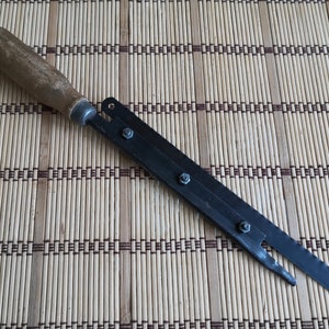 May include: A long, thin, black metal tool with a wooden handle. The tool has four screws along the metal blade and a pointed tip.
