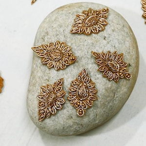 Könnte beinhalten: Eine Sammlung von acht goldfarbenen Metallembellishments in Form von Blättern und Tropfen. Die Verzierungen sind auf einem grauen Stein und einem weißen Hintergrund angeordnet.