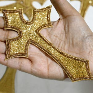 Puede incluir: Adornos dorados en forma de cruz, intrincadamente con cuentas y delineados con un hilo dorado más oscuro. Las cruces tienen un diseño detallado y se muestran sobre un fondo neutro. El texto "NAAP-1385" es visible en la esquina superior izquierda.