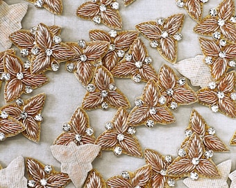 Op de afbeelding: Een verzameling van gouden en zilveren geborduurde bloemvormige versieringen met heldere strass steentjes. De versieringen zijn gerangschikt op een witte stoffen achtergrond.