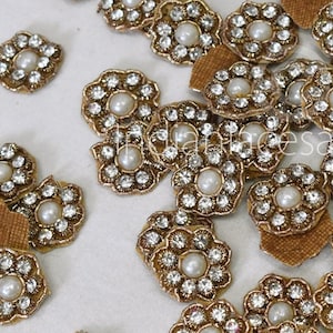 May include: A collection of gold-toned flower-shaped embellishments with clear rhinestones and a single white pearl in the center. The embellishments are scattered on a white surface.