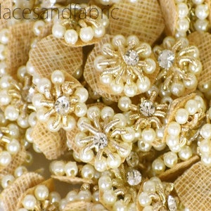May include: A pile of beige and gold fabric embellishments with pearl beads and rhinestones. The embellishments are shaped like flowers and have a burlap backing.