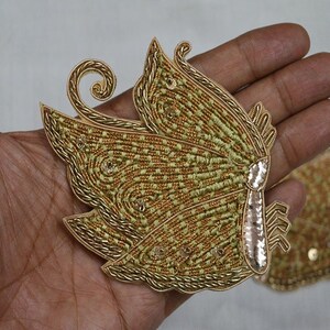 May include: Close-up of two gold butterfly appliques. The butterfly wings are intricately embroidered with gold thread and small sequins. The appliques have a light green and gold color scheme, with a detailed design.