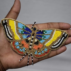 Pode incluir: Aplique de borboleta bordada com contas amarelas, azuis, laranjas e brancas. A borboleta tem um contorno dourado e é adornada com pequenas pérolas. As asas apresentam padrões detalhados e uma variedade de cores de contas.