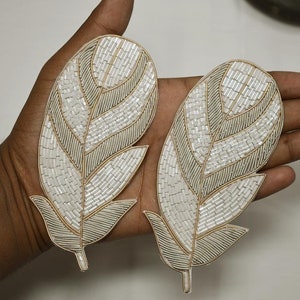 May include: Two white and gold beaded feather-shaped appliques. The appliques are made of small, white beads and gold thread. They are perfect for adding a touch of elegance to any project.