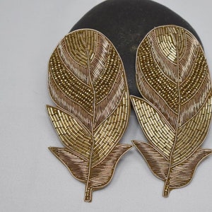 May include: Two gold and brown beaded feather-shaped embellishments. The feathers are made of small beads and have a detailed design.