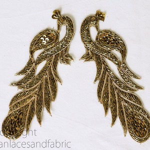 Könnte beinhalten: Zwei goldene Pfauenverzierungen mit Pailletten und Perlen. Die Pfauen sind in entgegengesetzte Richtungen gerichtet und haben ihre Schwänze ausgebreitet.