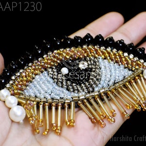 Puede incluir: Una aplicación de ojo de cuentas negras, doradas y blancas con una franja de cuentas doradas. El ojo está rodeado de cuentas de perlas blancas.