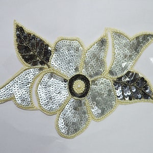 May include: A silver and black sequined flower applique with a pearl center. The applique is perfect for adding a touch of sparkle to clothing, bags, or home decor.
