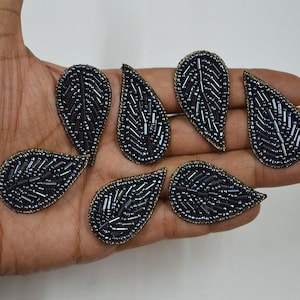 May include: Eight black beaded leaf-shaped embellishments with gold trim. The embellishments are arranged on a hand.